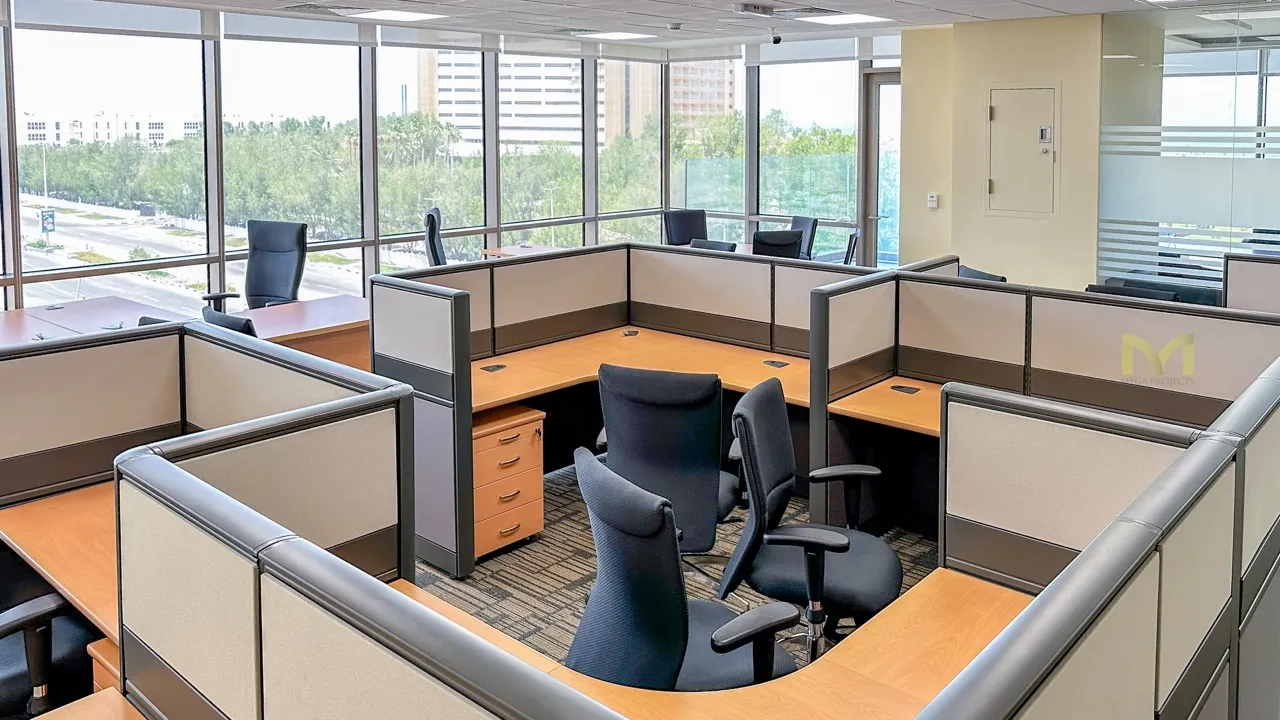 Modern office with cubicles, ergonomic chairs, large windows, and a view of trees and buildings outside.