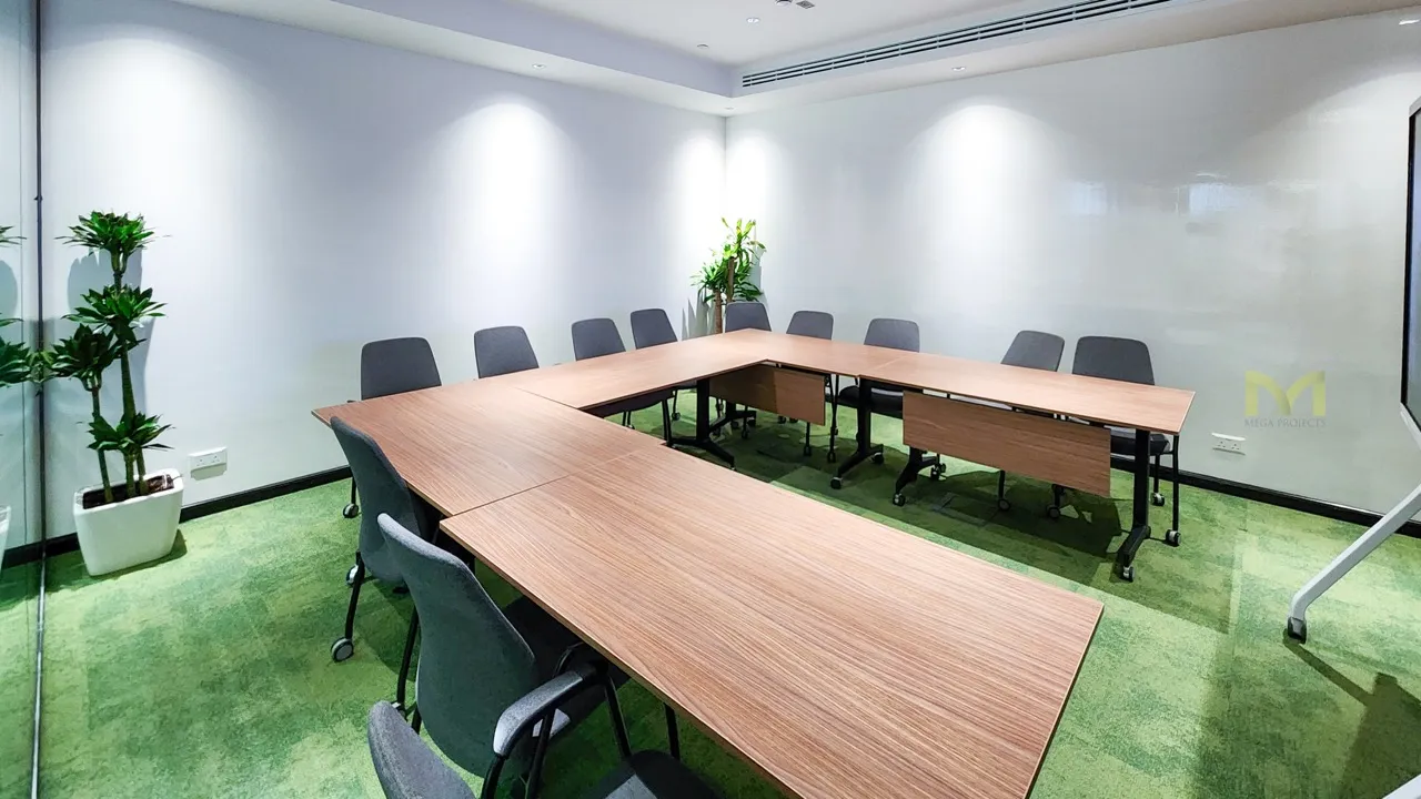 Modern conference room with U-shaped wooden tables, grey chairs, and green carpet flooring.