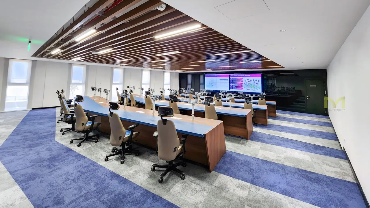 Modern conference room with rows of chairs, long blue tables, and large screens on the wall.