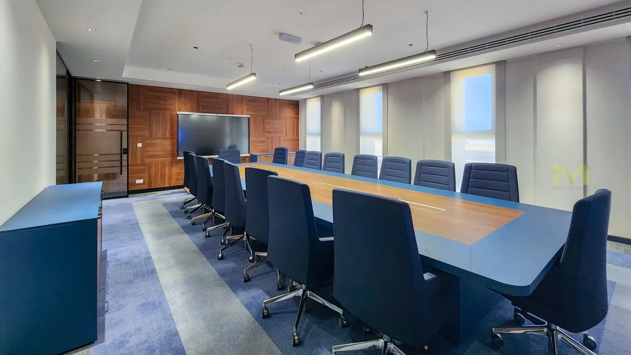 Modern conference room with long blue and wood table, blue chairs, and wall-mounted screen.