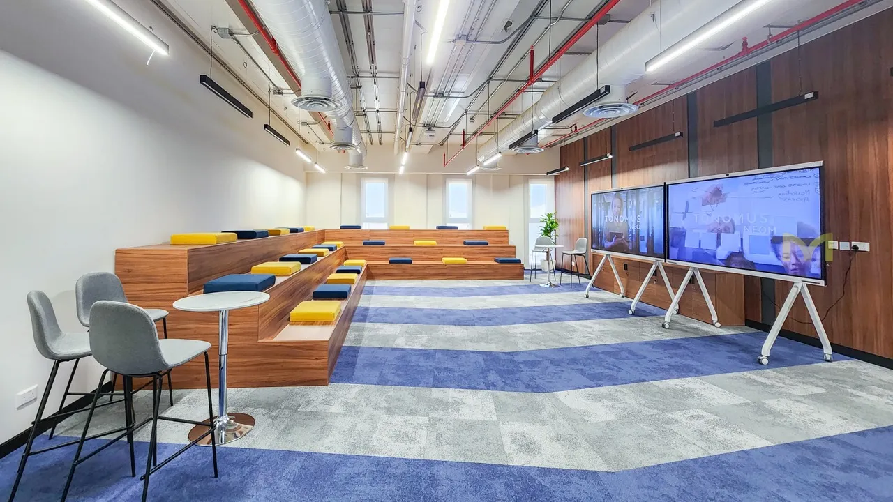 Modern conference room with tiered wooden seating, colorful cushions, two large screens, and gray chairs.