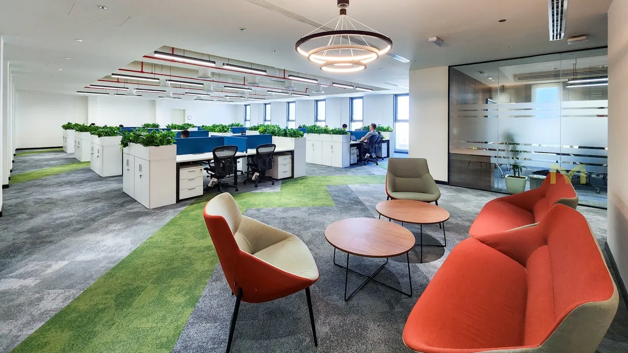 Modern office space with green and gray carpet, orange and gray chairs, round tables, and workstations with plants.
