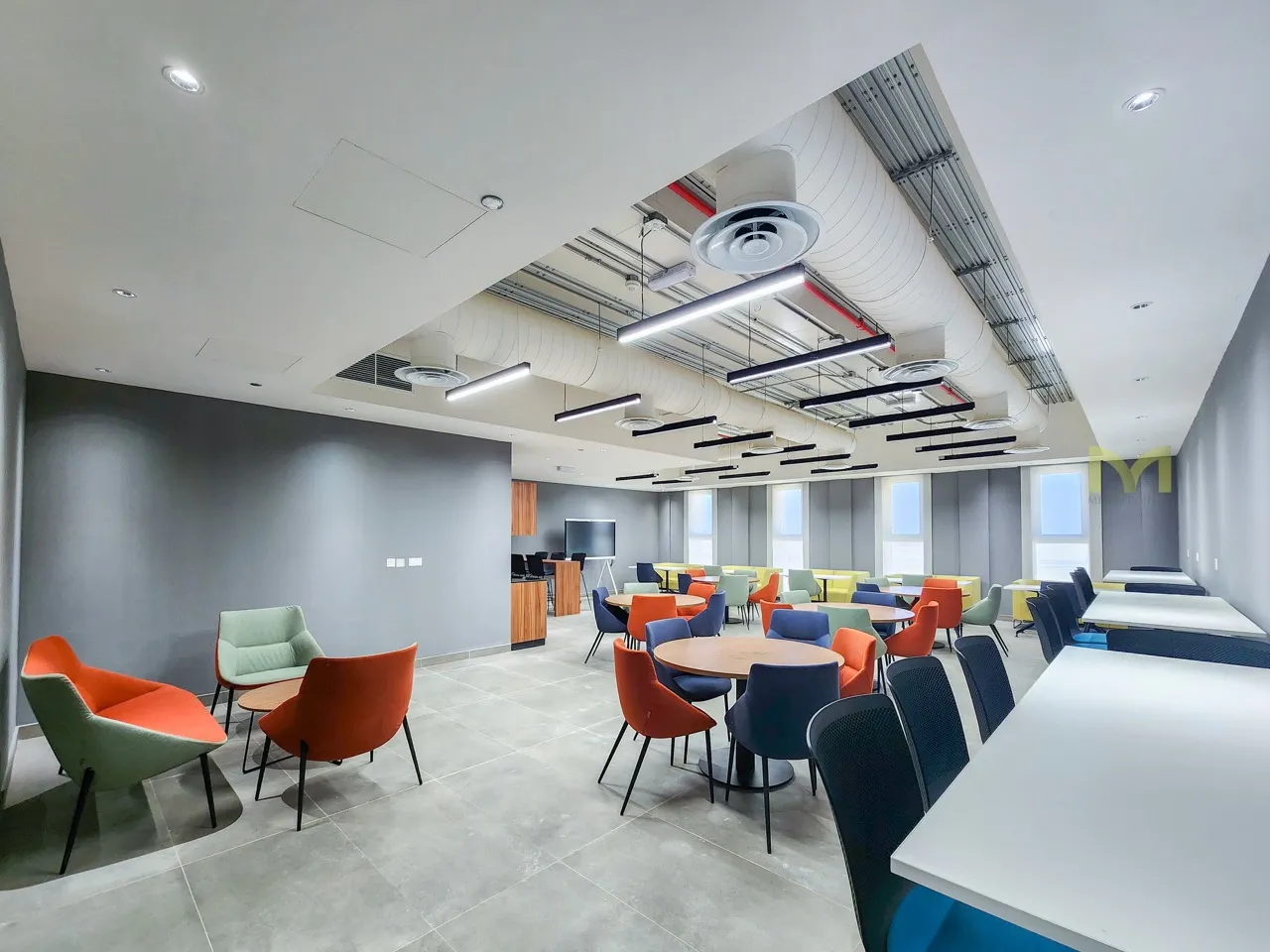 Modern office lounge with colorful chairs, round tables, desks, and large windows under white ceiling.