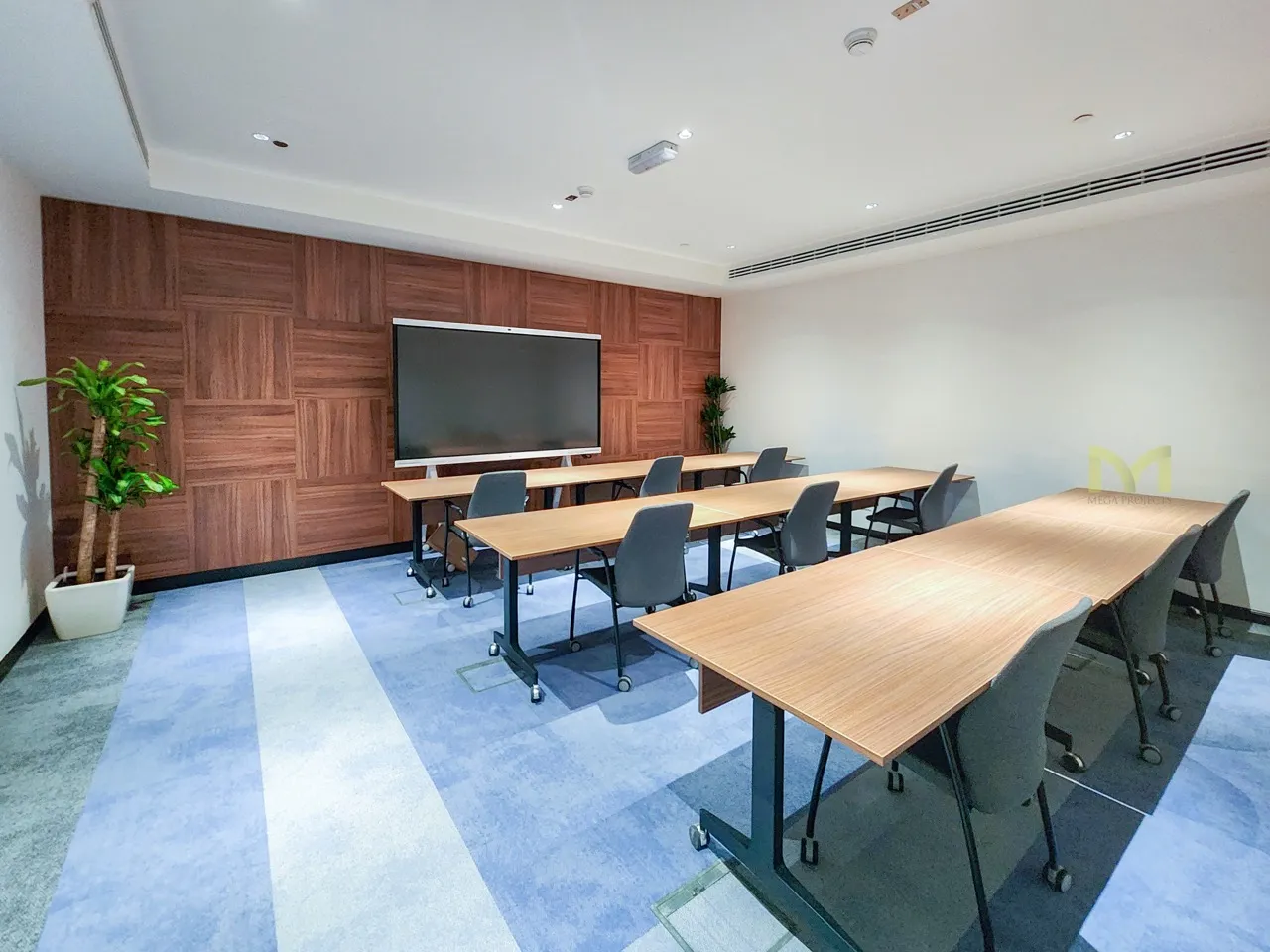 Modern conference room with wooden tables, black chairs, a large screen, and potted plants.