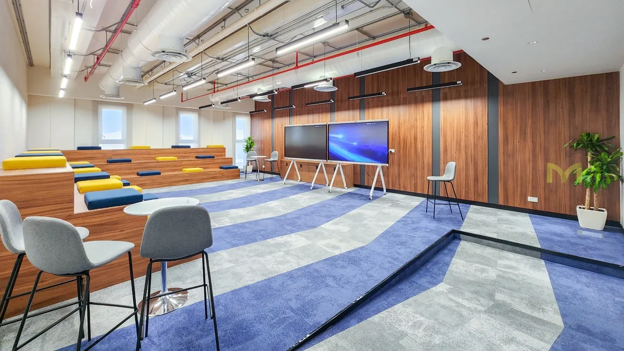 Modern conference room with tiered seating, blue-gray carpet, wooden walls, and two large screens.