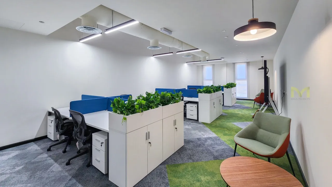 Modern office with white desks, blue dividers, black chairs, green plants, and a seating area with armchairs.