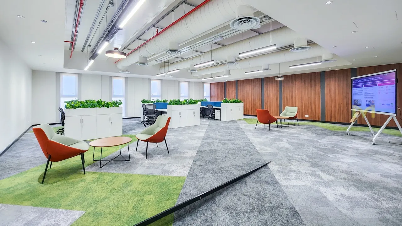 Modern office space with chairs, workstations, plants, wood-paneled wall, and a presentation screen.