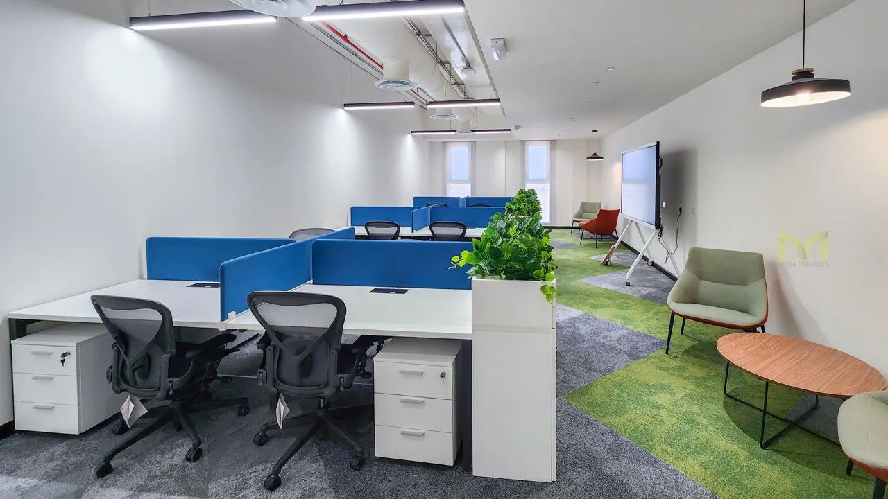Modern office workspace with white desks, blue dividers, black chairs, plants, and a seating area with chairs and table.