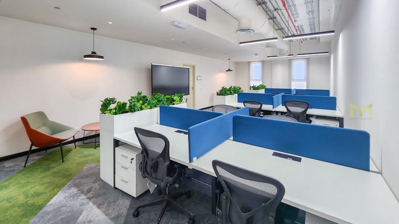 Modern office with white desks, blue dividers, black chairs, green plants, and lounge chair on carpet.