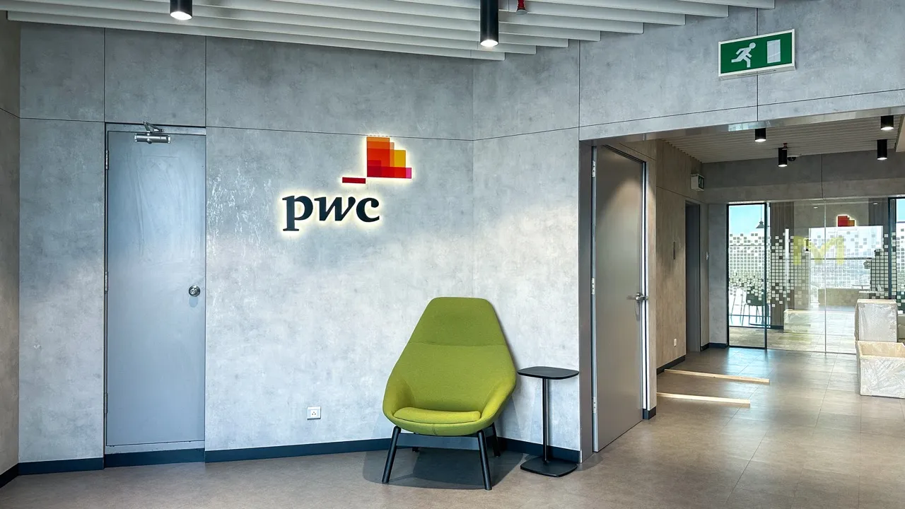 Modern office corner with gray walls, green chair, small table, and illuminated PwC logo on wall.