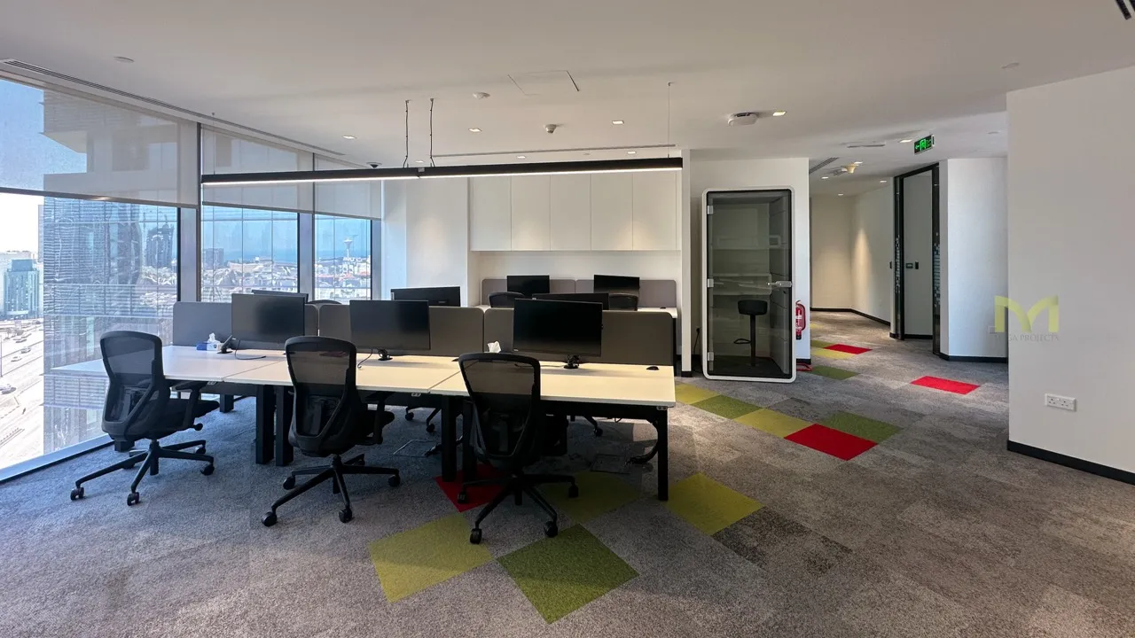 Modern office with desks, black chairs, computers, and large windows showing city view.