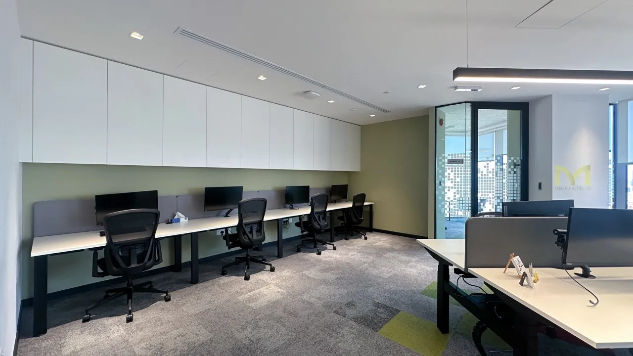 Modern office space with desks, computer monitors, ergonomic chairs, and glass door to a meeting room.