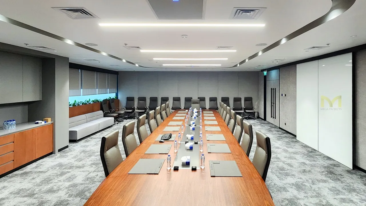 Modern boardroom with long wooden table, leather chairs, water bottles, and gray carpeted floor.