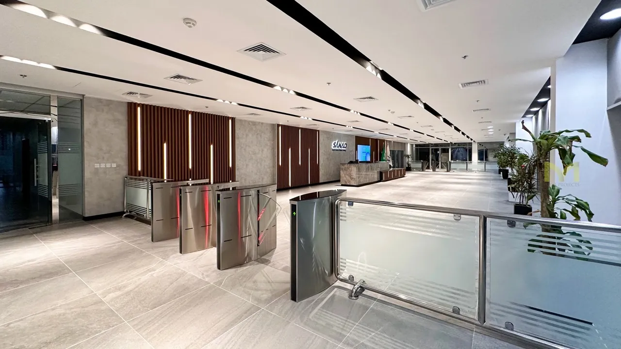 Modern office lobby with turnstiles, reception desk, and decorative plants under bright ceiling lights.