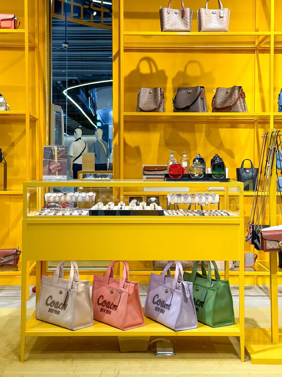 Yellow retail display with purses, watches, perfume bottles, and four colorful Coach bags on a bottom shelf.