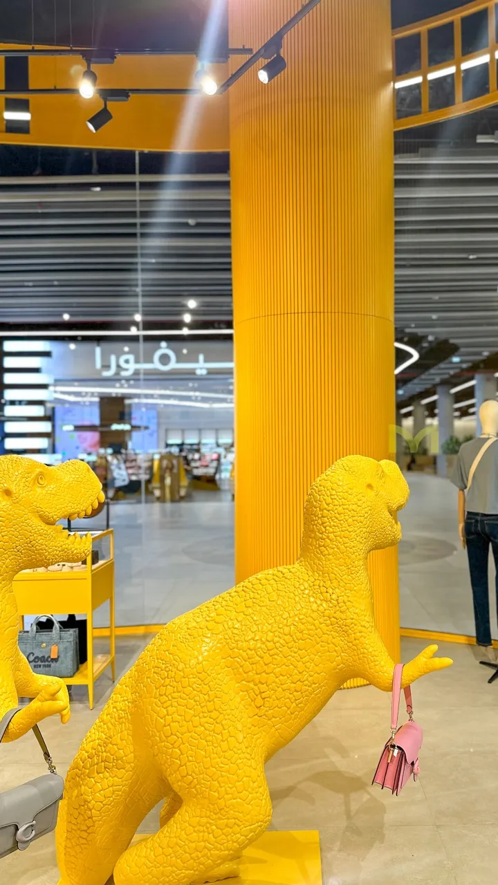Yellow dinosaur statues holding handbags in a brightly lit retail store with mannequins.