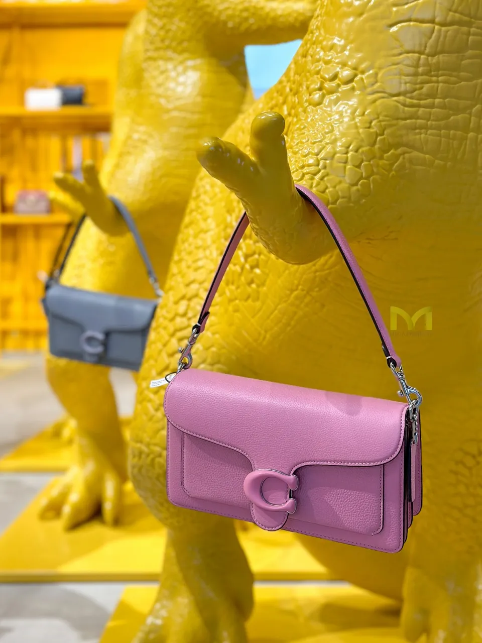 Pink leather handbag hanging from the claw of a yellow dinosaur statue in a retail display.