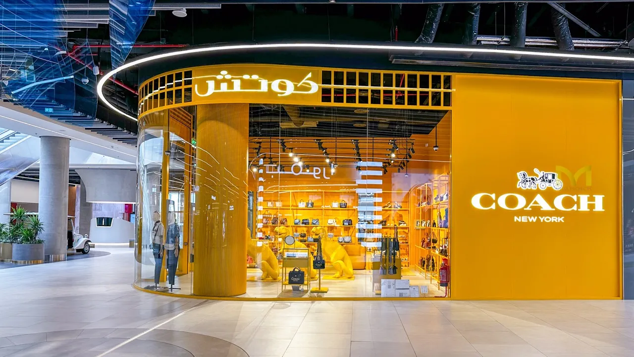 Bright yellow Coach New York store with glass walls, handbags on shelves, and large dog statues inside.