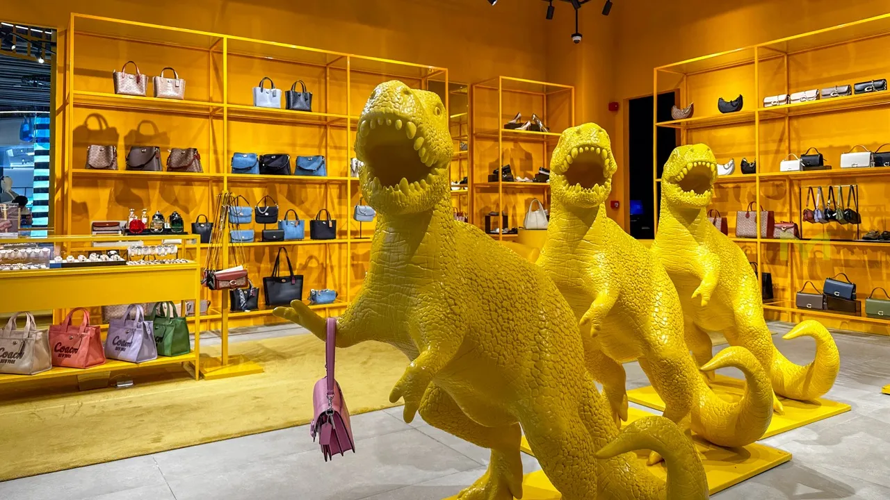 Bright yellow dinosaur statues holding handbags in a colorful retail store with shelves of purses.