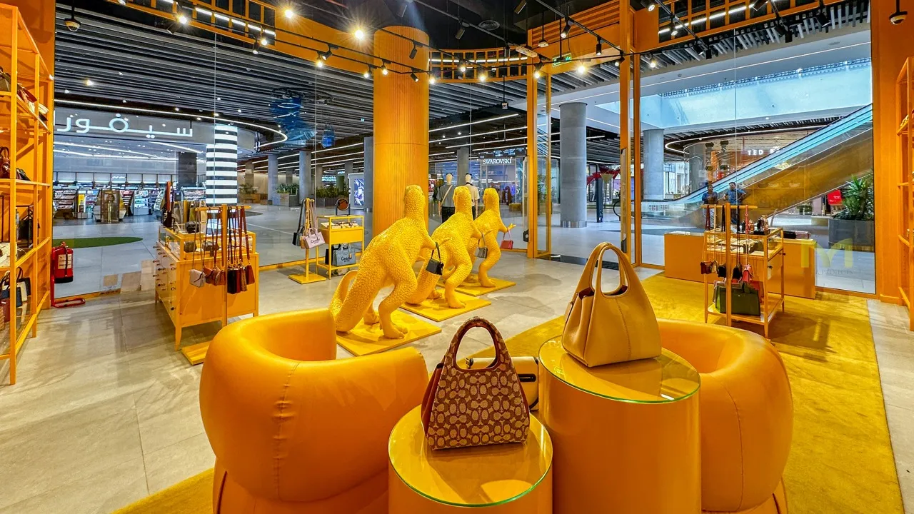 Bright yellow retail space featuring plush chairs, handbags, and dinosaur statues holding bags.