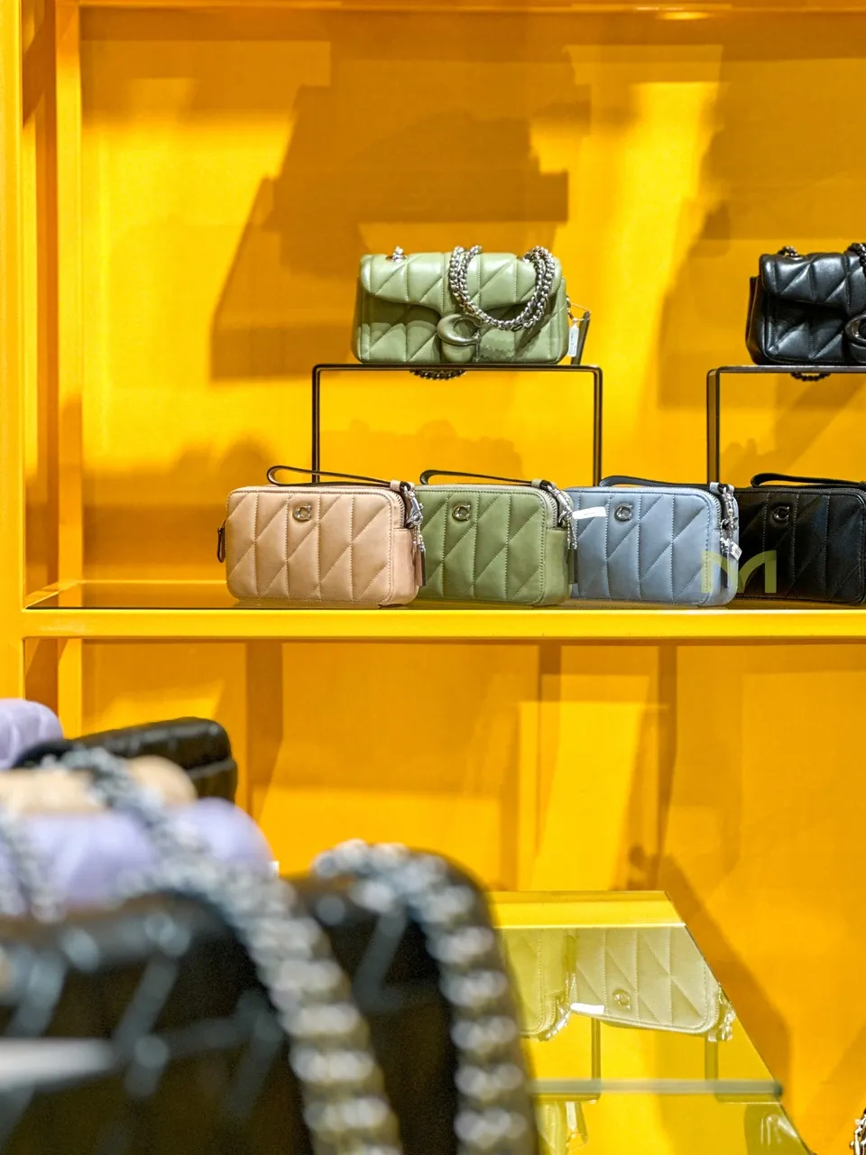Display of five quilted handbags in beige, green, blue, and black on yellow shelves in a store.