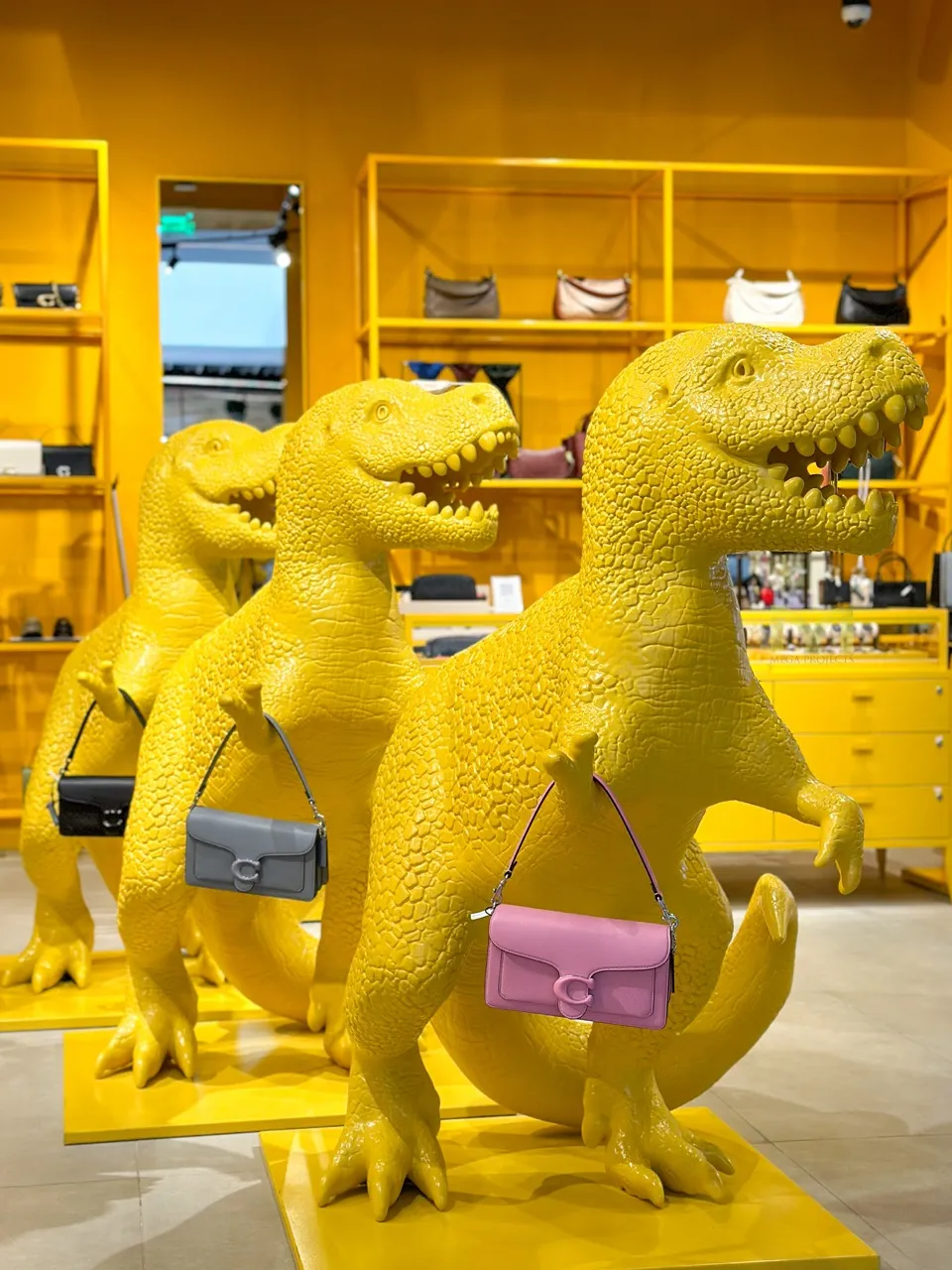 Three large yellow dinosaur statues holding different colored handbags in a bright retail store.
