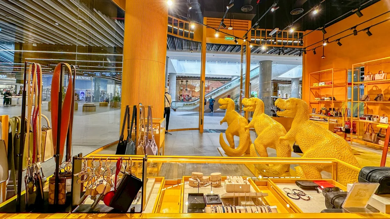 Retail space with orange walls, dinosaur statues, handbags, and jewelry displays near mall escalators.