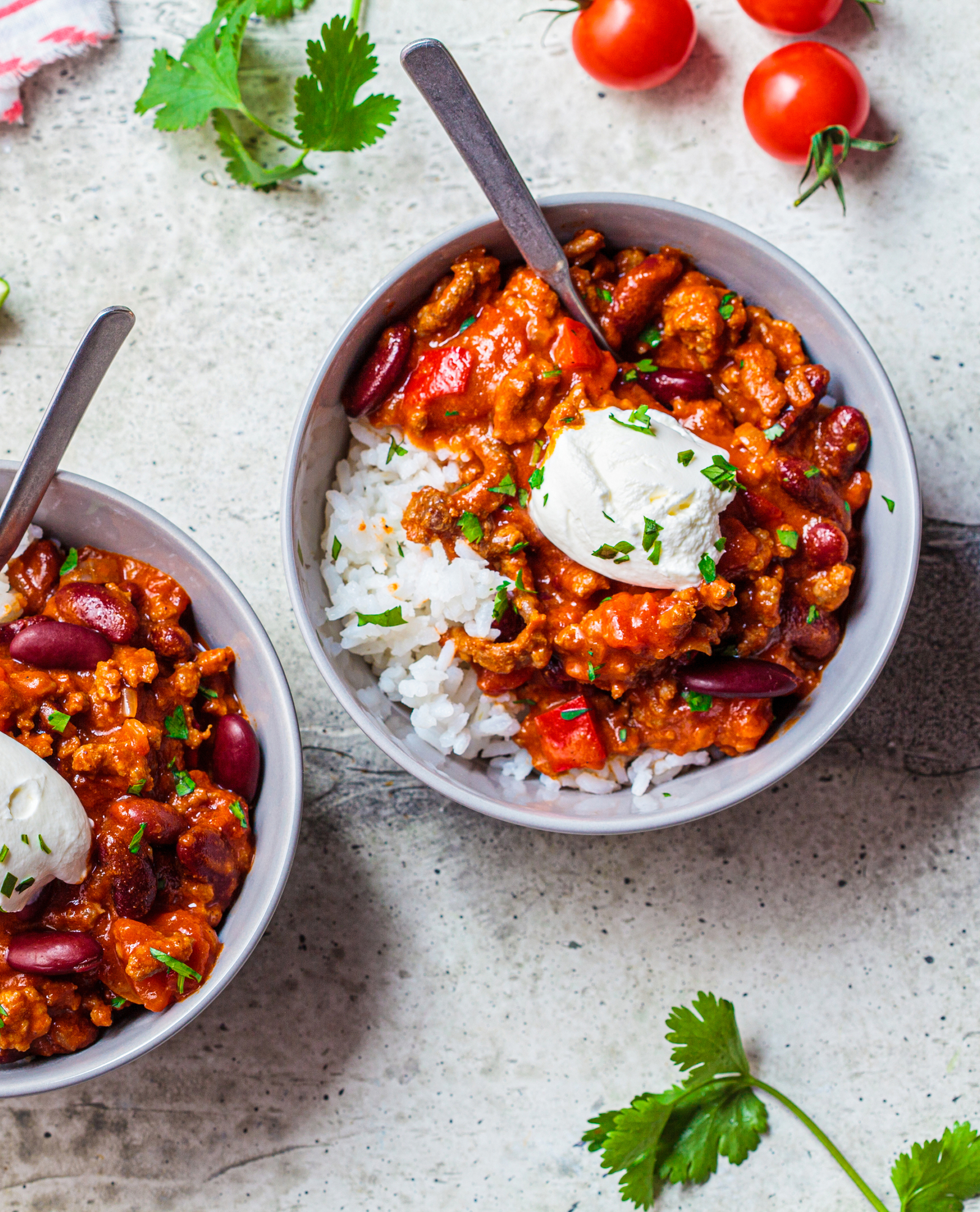 Deux bols de chili con carne avec riz, garnis de crème aigre et coriandre fraîche, à côté de tomates cerises et feuilles de coriandre.