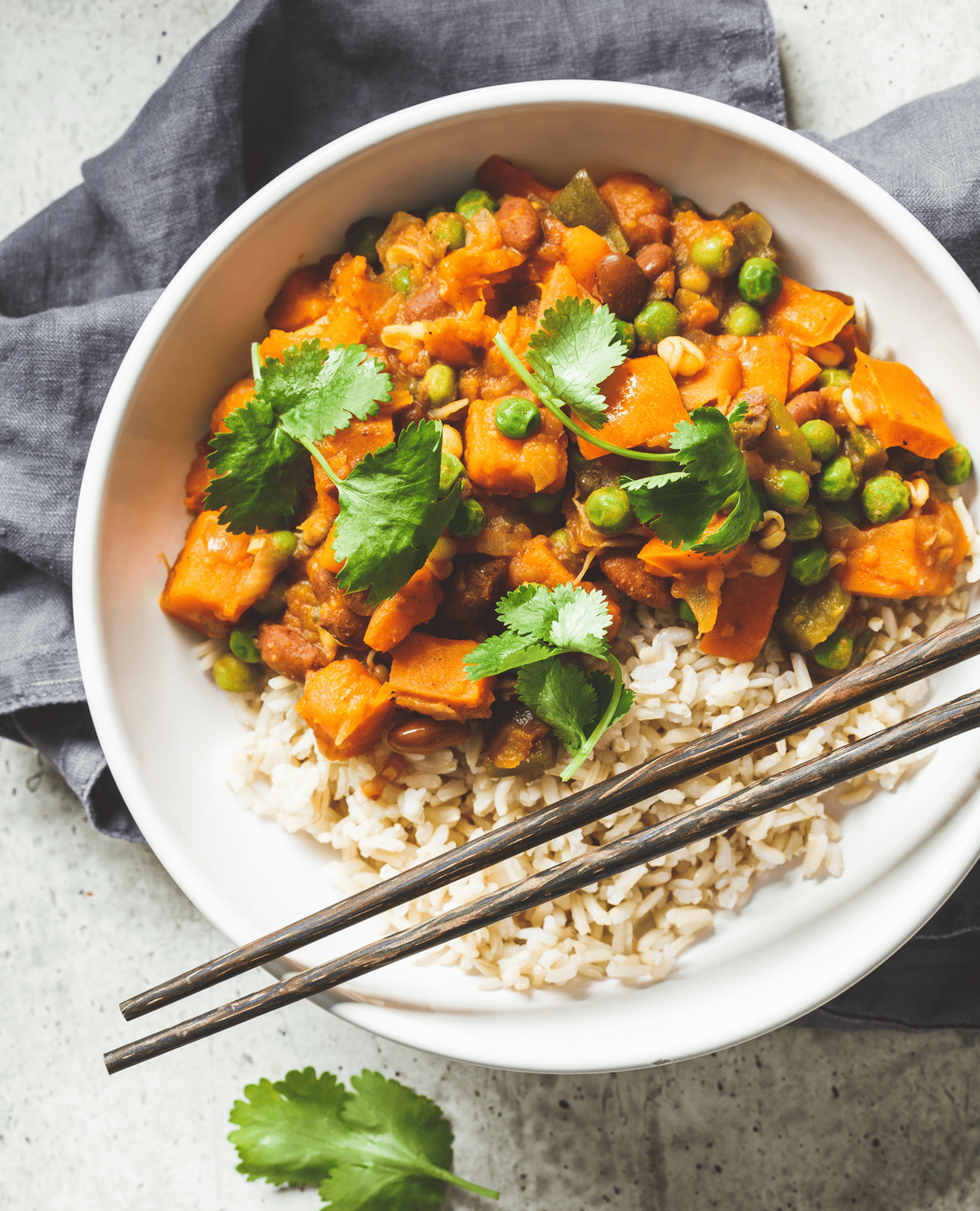 Bol de riz brun garni d'un curry de légumes coloré avec pois verts, patates douces, haricots rouges et coriandre fraîche, accompagné de baguettes en bois.