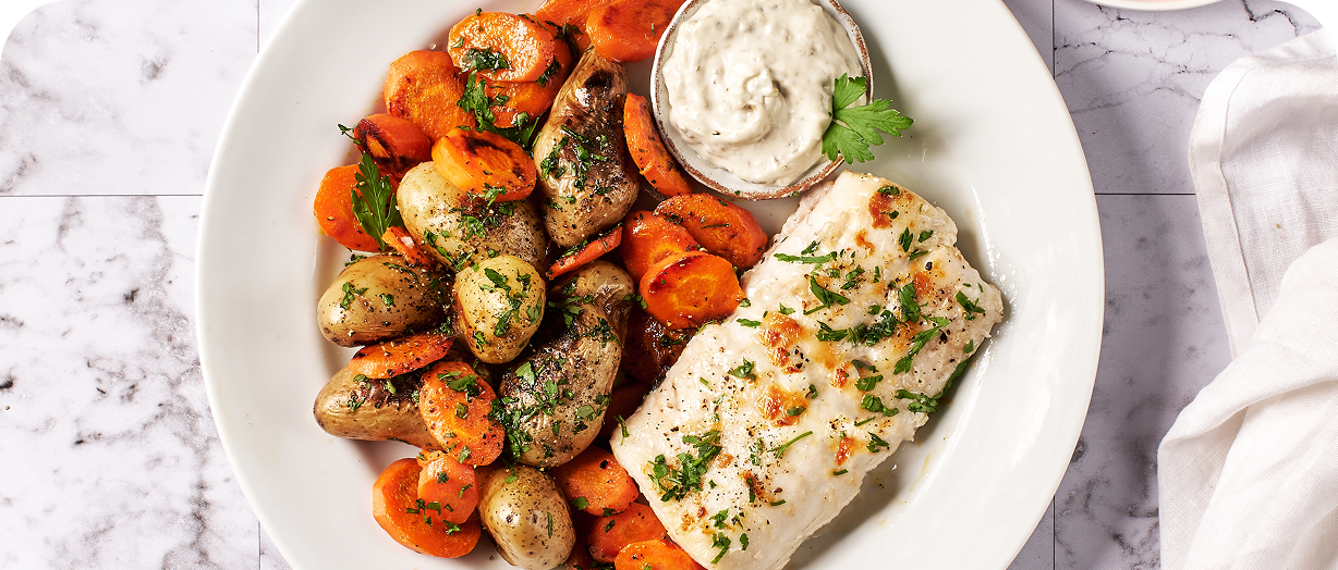 Filet de poisson grillé avec pommes de terre rattes, carottes sautées et sauce blanche dans une assiette blanche.