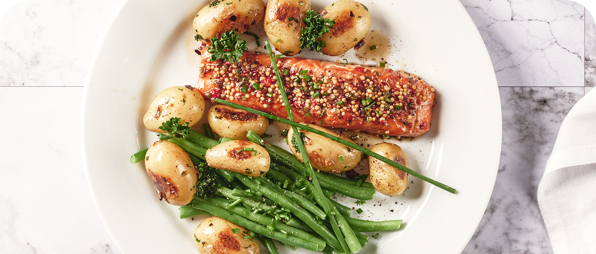 Filet de saumon grillé avec graines de moutarde, accompagné de pommes de terre rôties et haricots verts sur une assiette blanche.