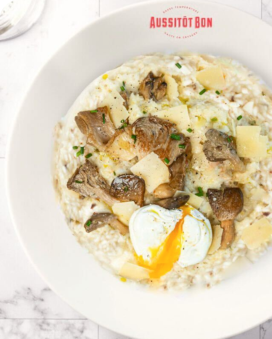 Risotto crémeux aux champignons, copeaux de fromage et œuf poché coulant dans une assiette blanche.