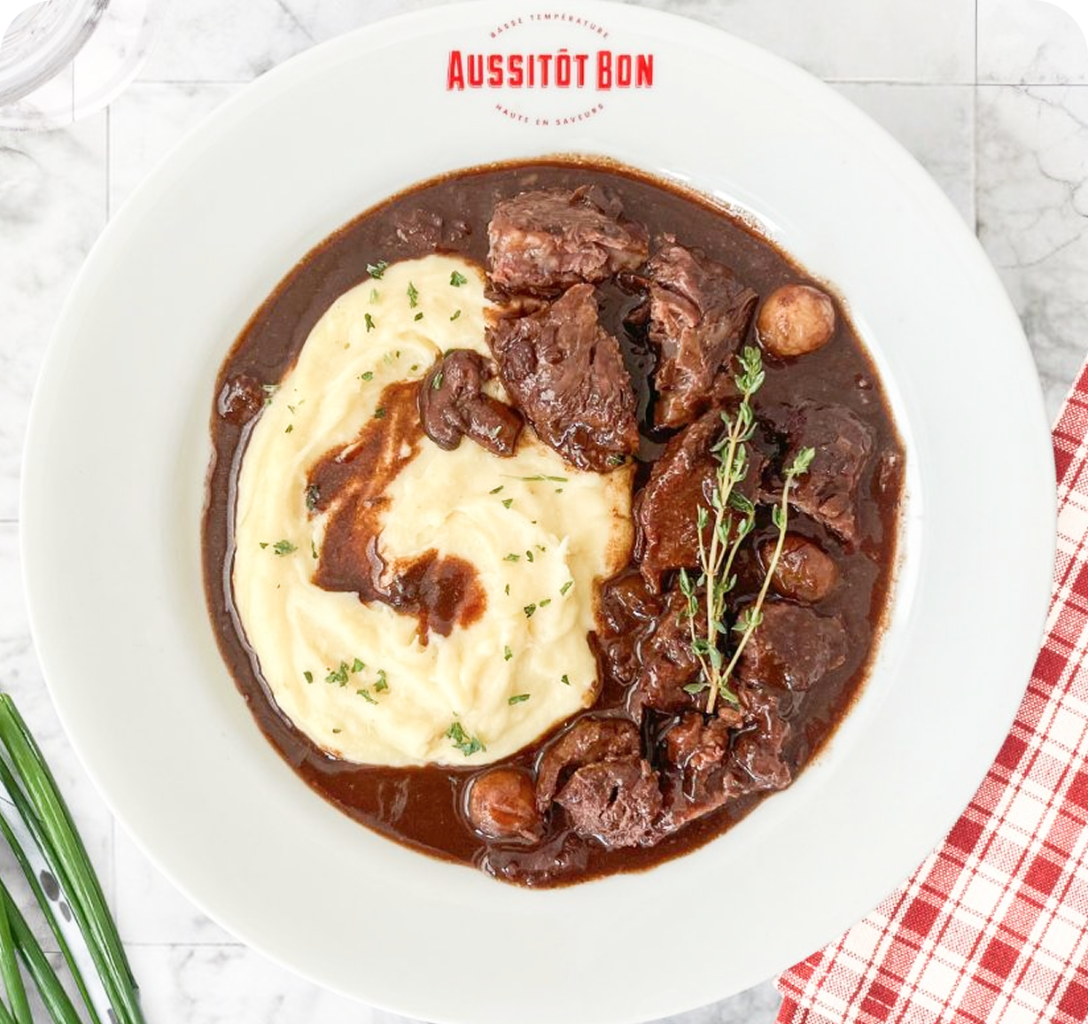 Plat de bœuf bourguignon avec purée de pommes de terre, garniture de thym, servie dans une assiette blanche avec le logo Aussitôt Bon.
