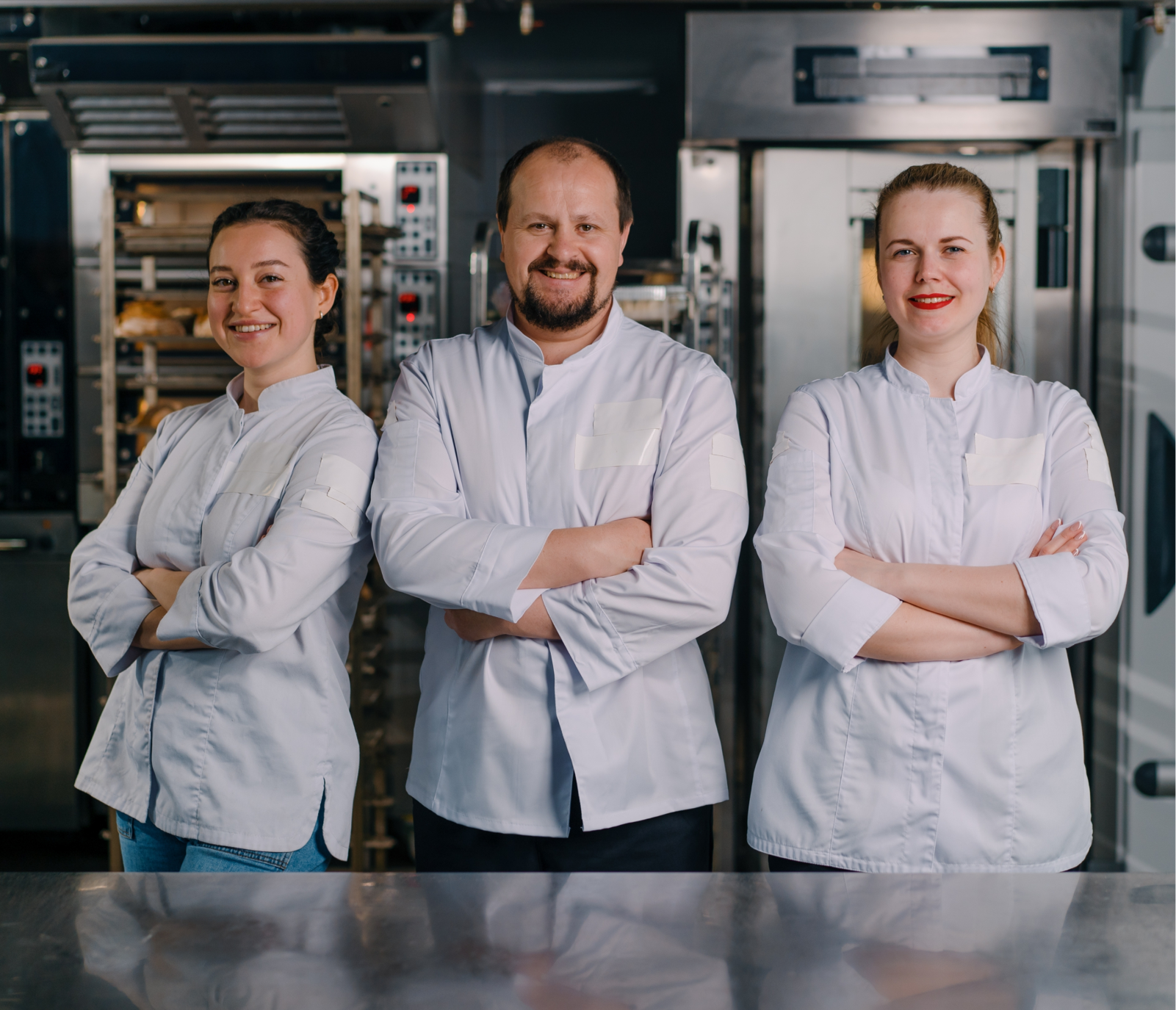 Trois boulangers souriants portant des vestes blanches avec les bras croisés dans une cuisine industrielle.
