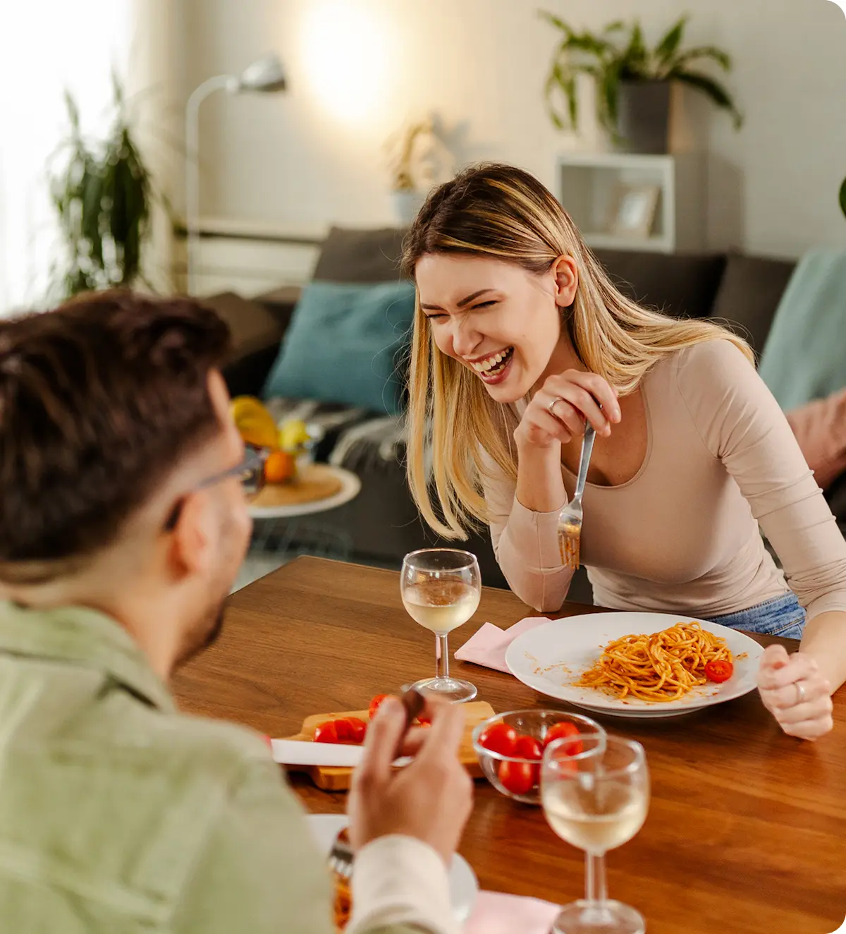 Un couple souriant partage un repas de spaghetti avec du vin blanc à une table en bois dans un salon moderne.