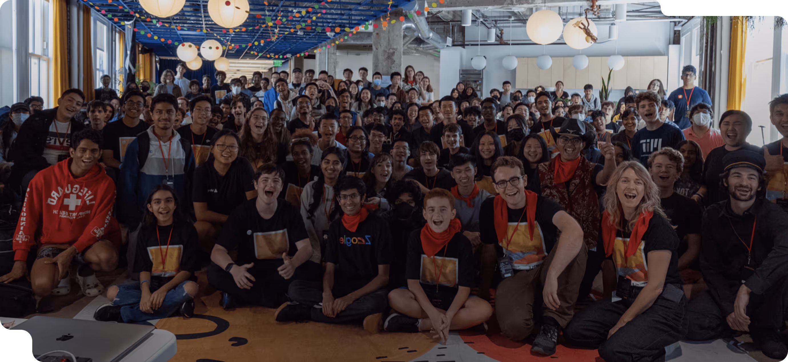Large diverse group of smiling young people posing together in a decorated indoor space with hanging lights and paper lanterns.