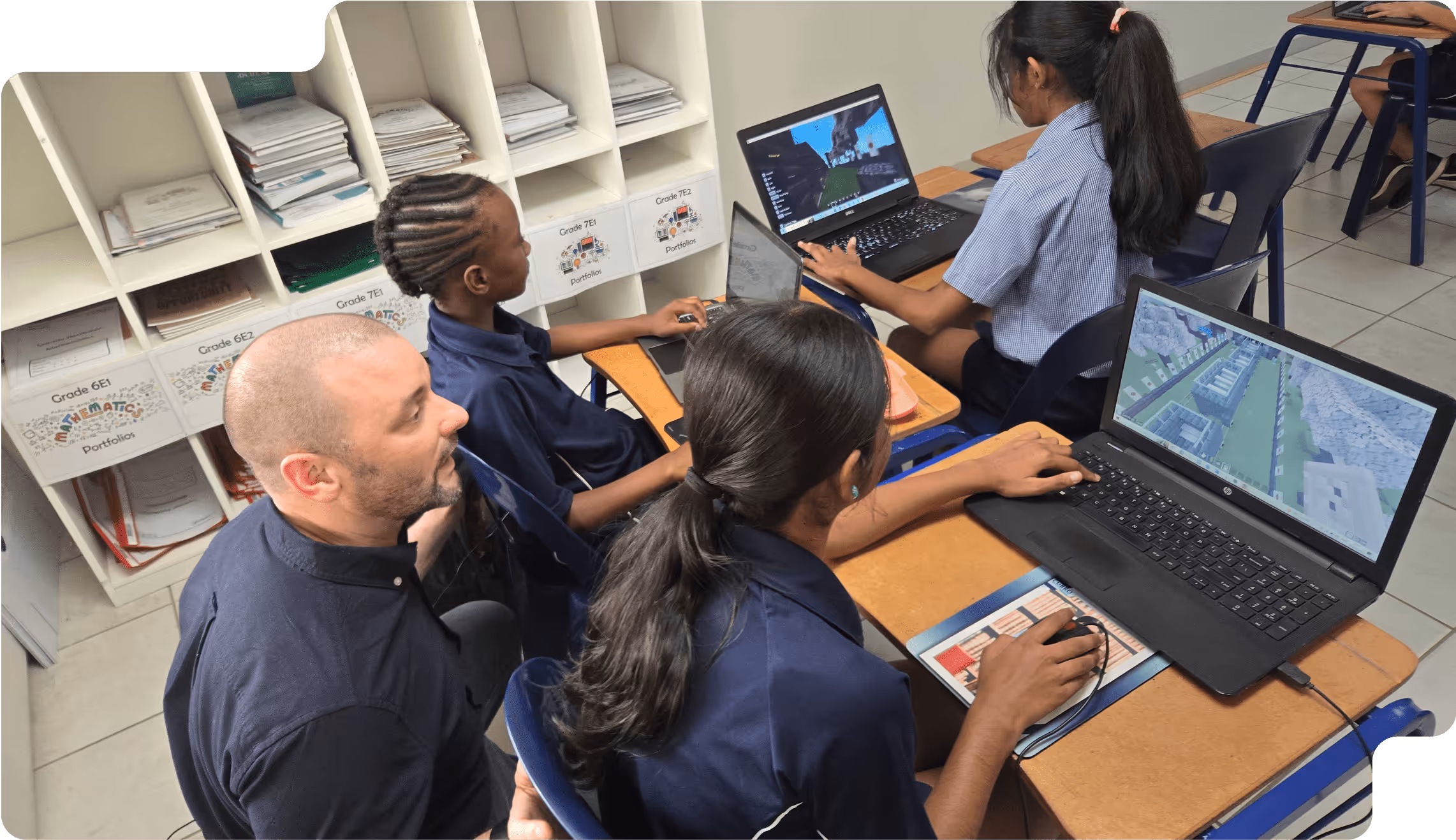Teacher assisting three students working on laptops in a classroom with papers organized on shelves behind them.