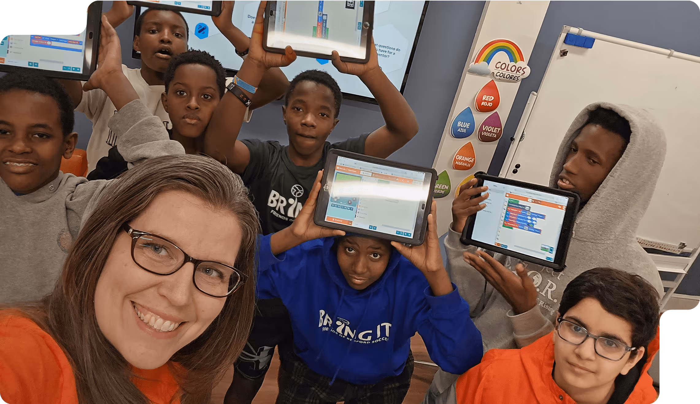 Smiling woman with glasses takes a selfie with a group of children holding tablets displaying coding programs in a classroom.