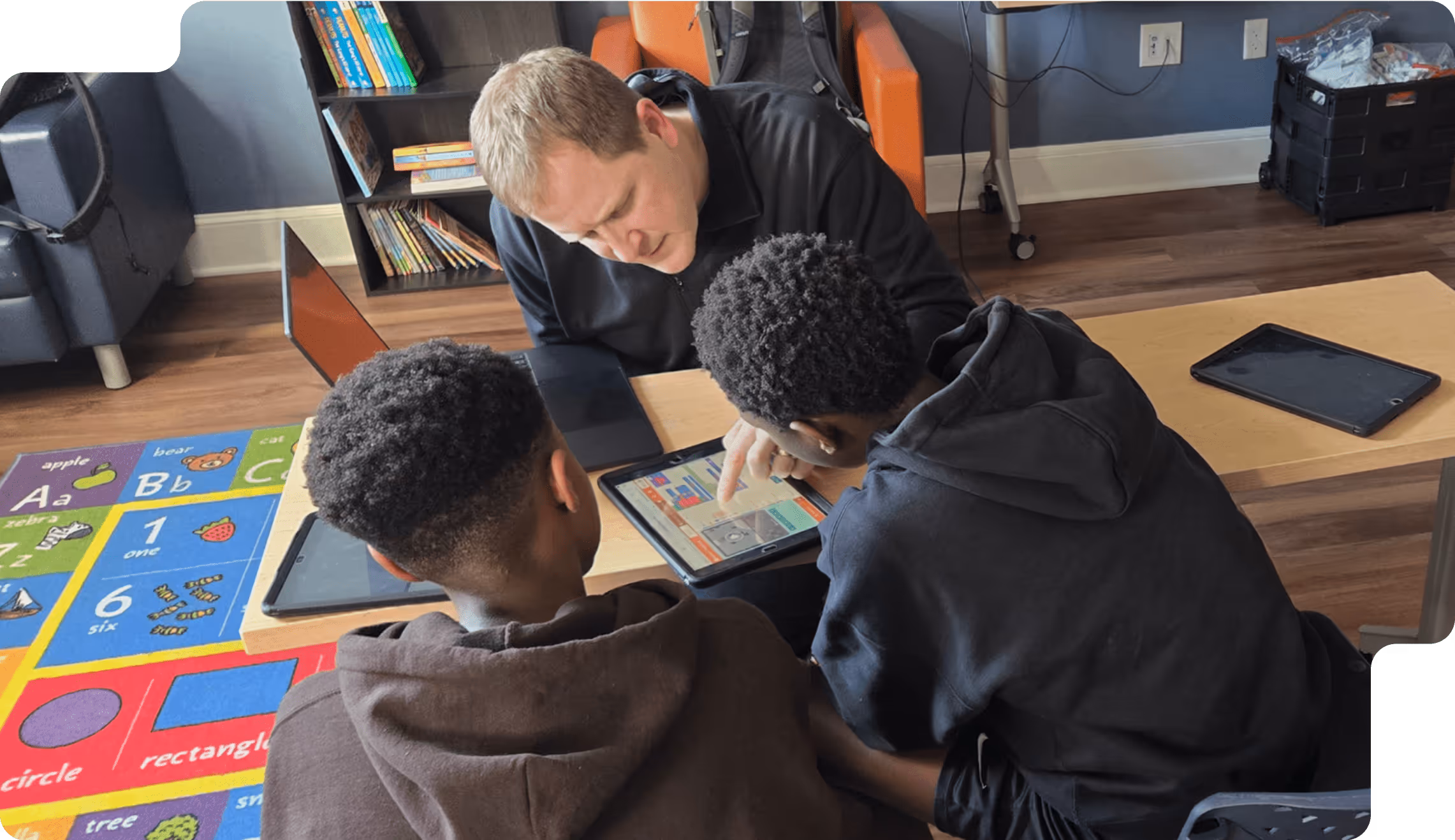 An adult male is assisting two boys using a tablet at a wooden table in a classroom.