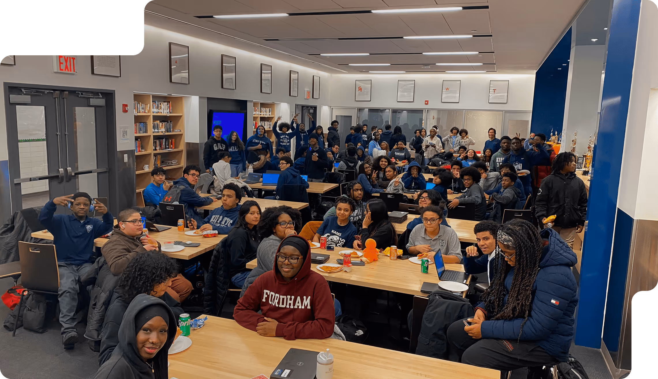A large group of diverse high school students sitting and standing in a library or classroom setting with laptops, snacks, and drinks on tables.