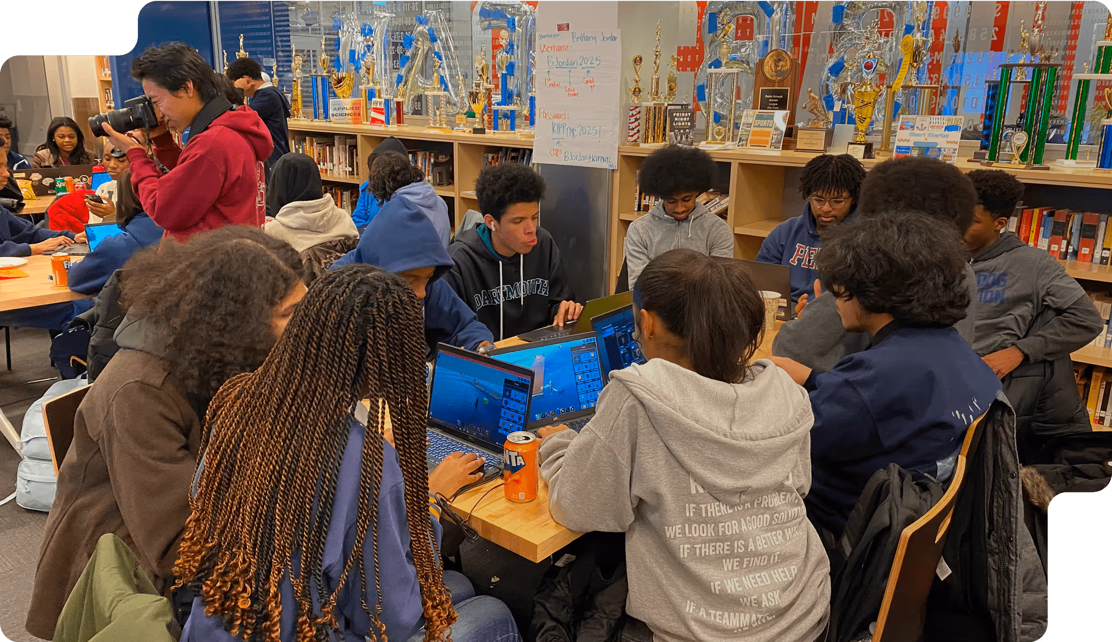 Group of teenagers working on laptops in a library with trophies displayed on shelves behind them and one person using a camera.