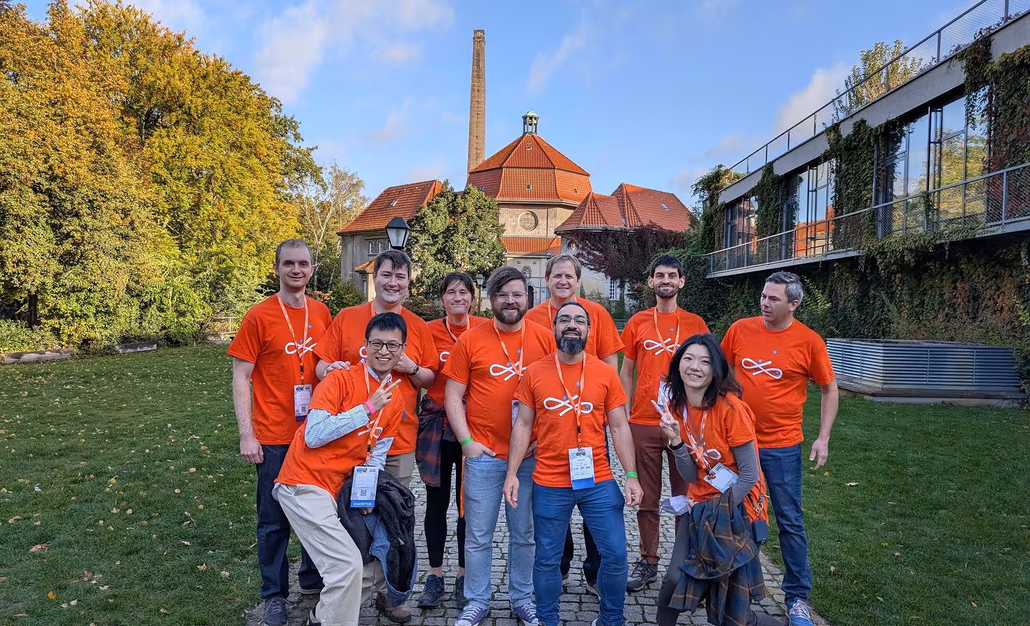 Group of ten people wearing matching orange t-shirts posing outdoors in front of a building with a red roof and surrounded by greenery.