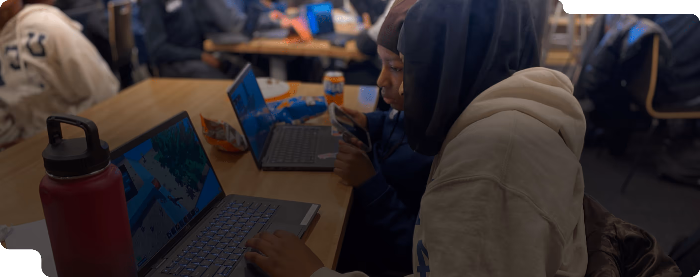 Two people sitting closely at a table working together on laptops, with a red water bottle and snack bags nearby.