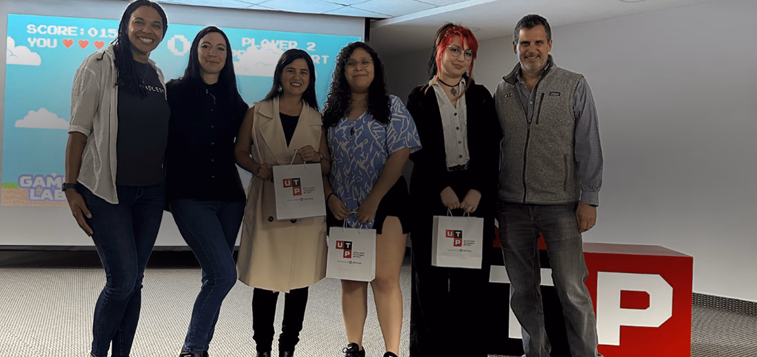 Group of six people standing indoors, three holding white bags with UTP logos, with a retro video game projected on a screen behind them.