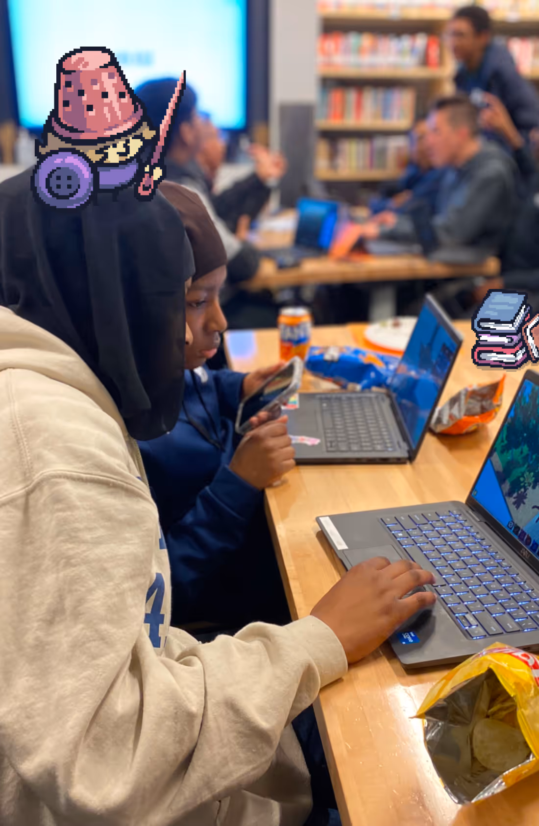 Young people working on laptops and mobile phones at a table with snacks in a library setting.