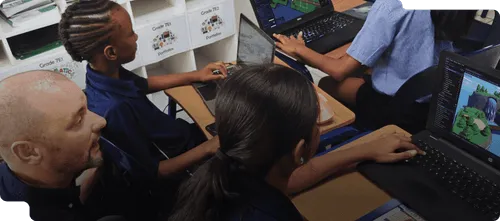 Teacher guiding students working on laptops, programming a 3D environment in a classroom.