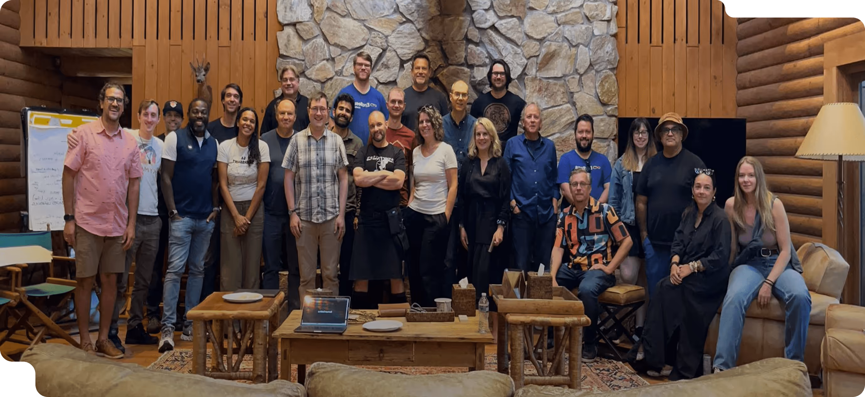 Group of diverse people standing and sitting in a cozy room with wooden walls and stone fireplace.