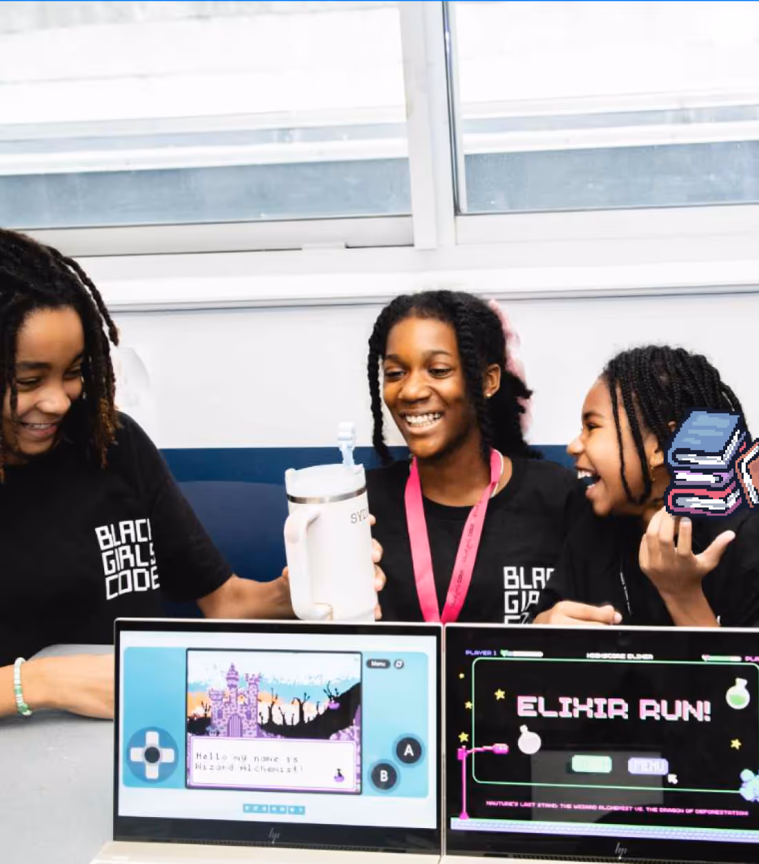 Three girls in Black Girls Code shirts smiling and interacting behind two laptops displaying pixel-art style games.