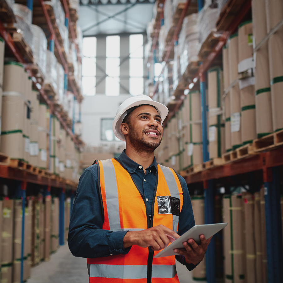 Warehouse worker wearing a white hard hat and orange safety vest, holding and using a tablet while standing between shelves stacked with large wrapped packages.