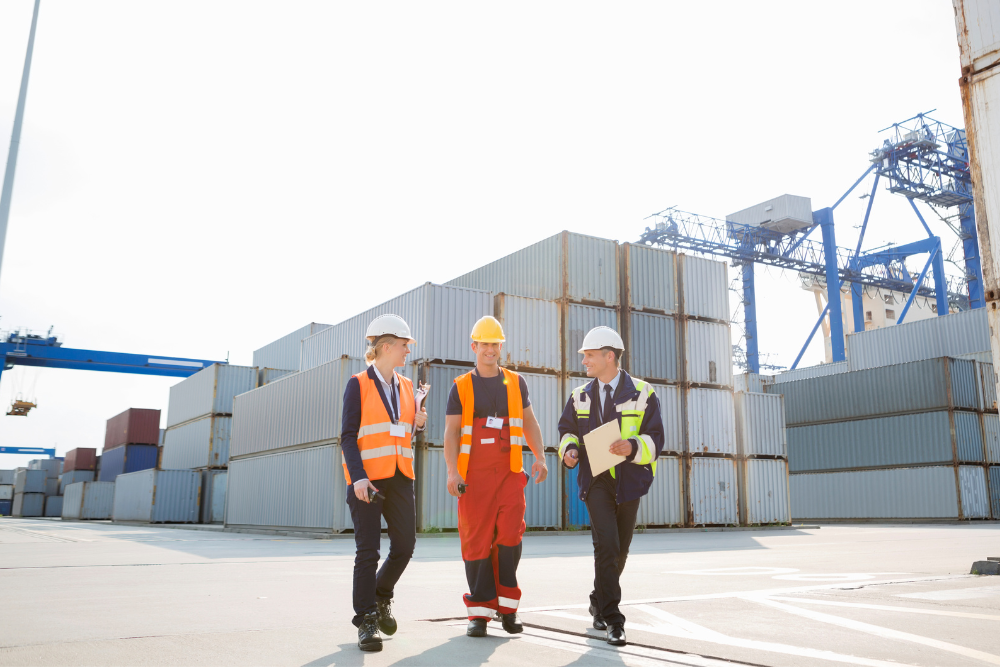 Three workers wearing safety helmets and vests walking and talking in front of stacked shipping containers at a port.